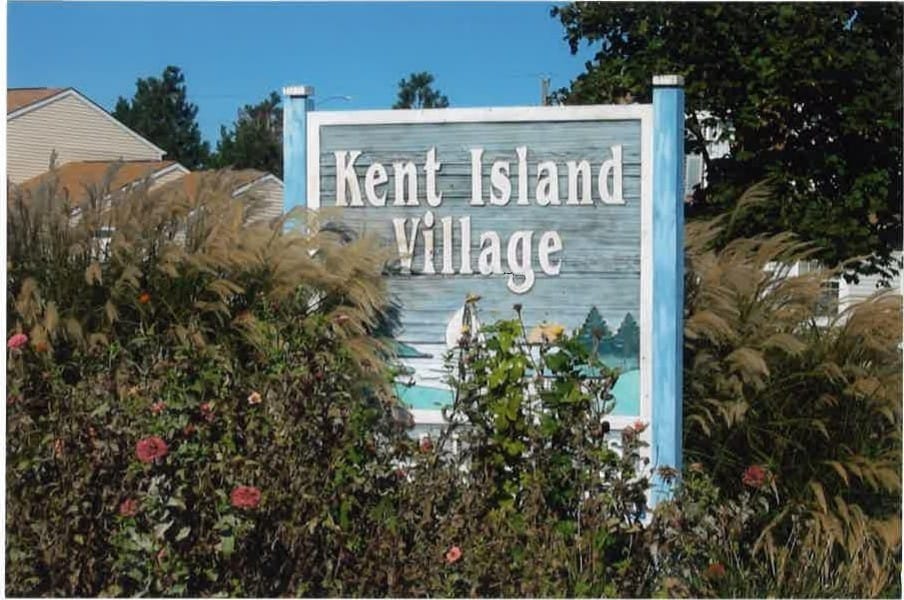 The Kent Island Village sign among ornamental grasses and flowers, with townhomes behind